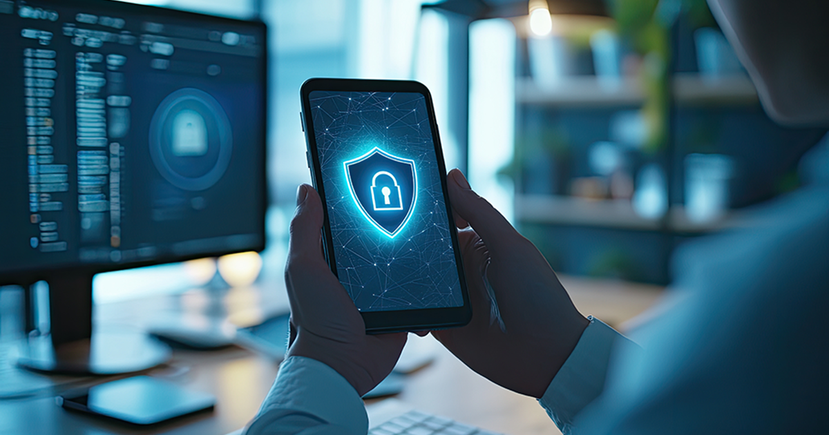 Person holding a smartphone displaying a glowing security shield icon, symbolizing mobile cybersecurity, with computer monitors in the background.