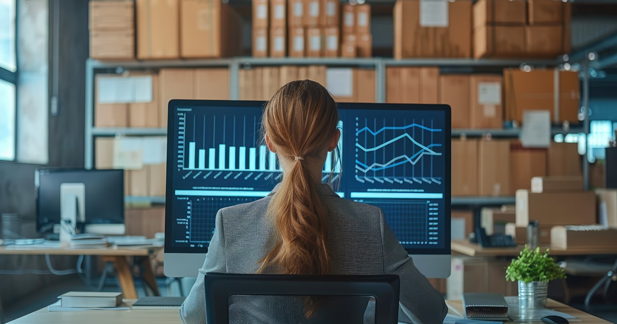 woman sitting in warehouse with boxes working large computer with graphs desplayed